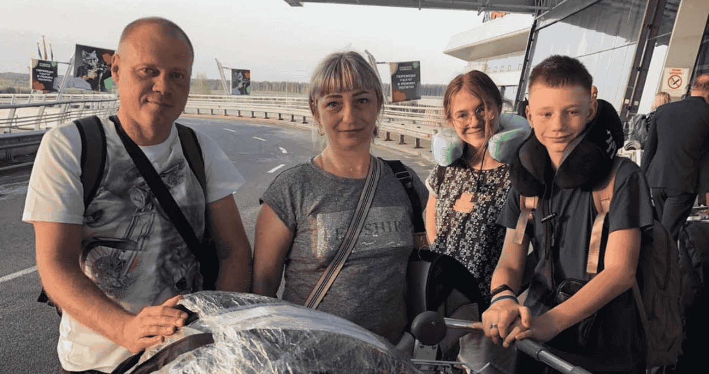 Семья из четырёх человек с багажом стоит у входа в аэропорт, готовясь к поездке; у детей подушки для шеи.