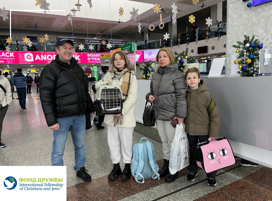 Семья из четырёх человек стоит в аэропорту с багажом и клеткой для домашнего животного; вокруг праздничные украшения и стойки регистрации.