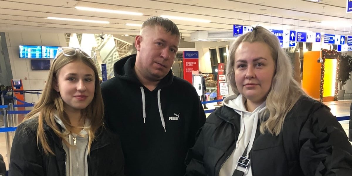 Семья из трёх человек — мужчина, женщина и девушка-подросток — стоят в аэропорту с багажом на фоне стоек регистрации.