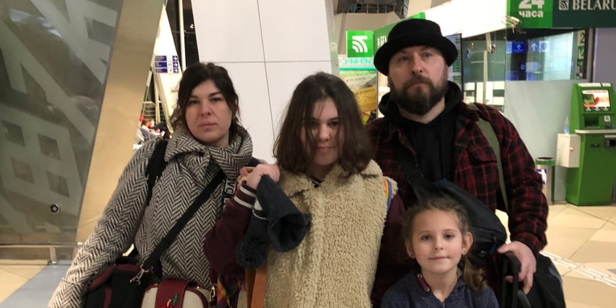 Семья из четырёх человек — мужчина, женщина и две девочки — стоят в аэропорту с сумками и багажом.