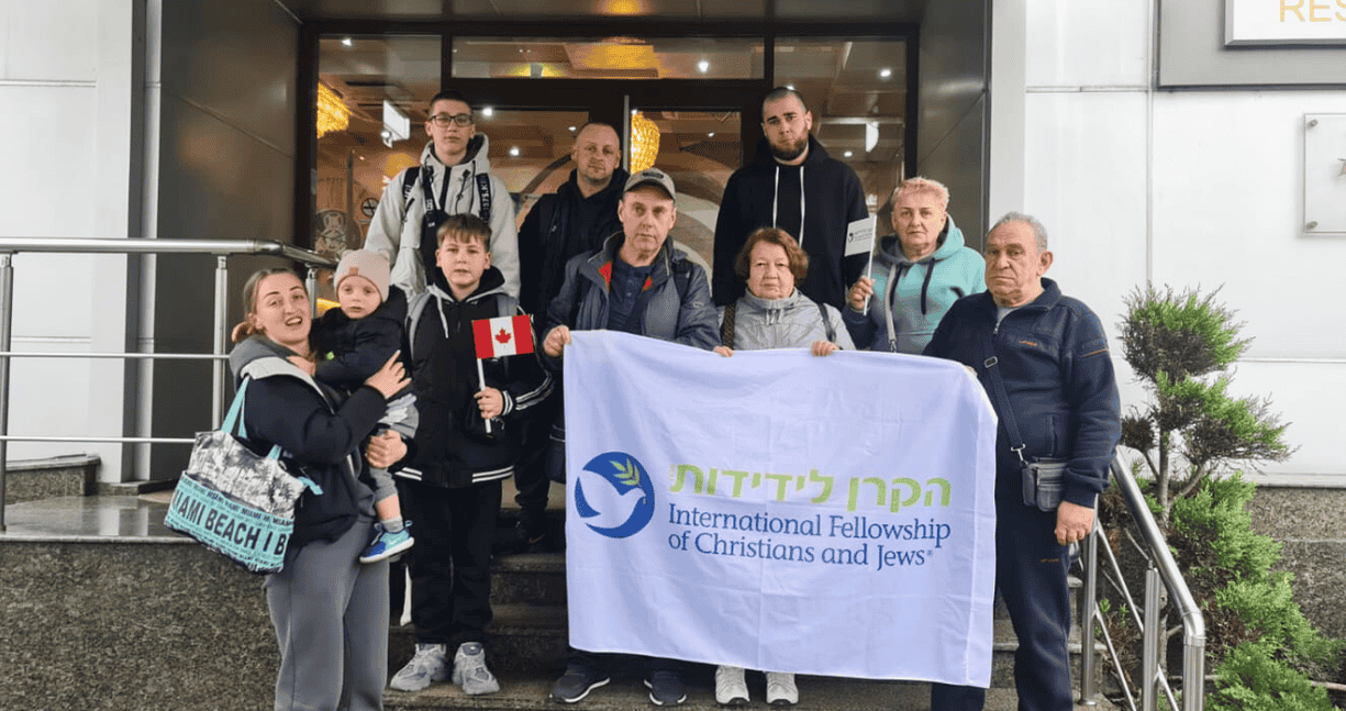 Группа людей стоит у входа в здание, держа флаг с логотипом Международного фонда христиан и евреев; один ребенок держит флаг Канады.