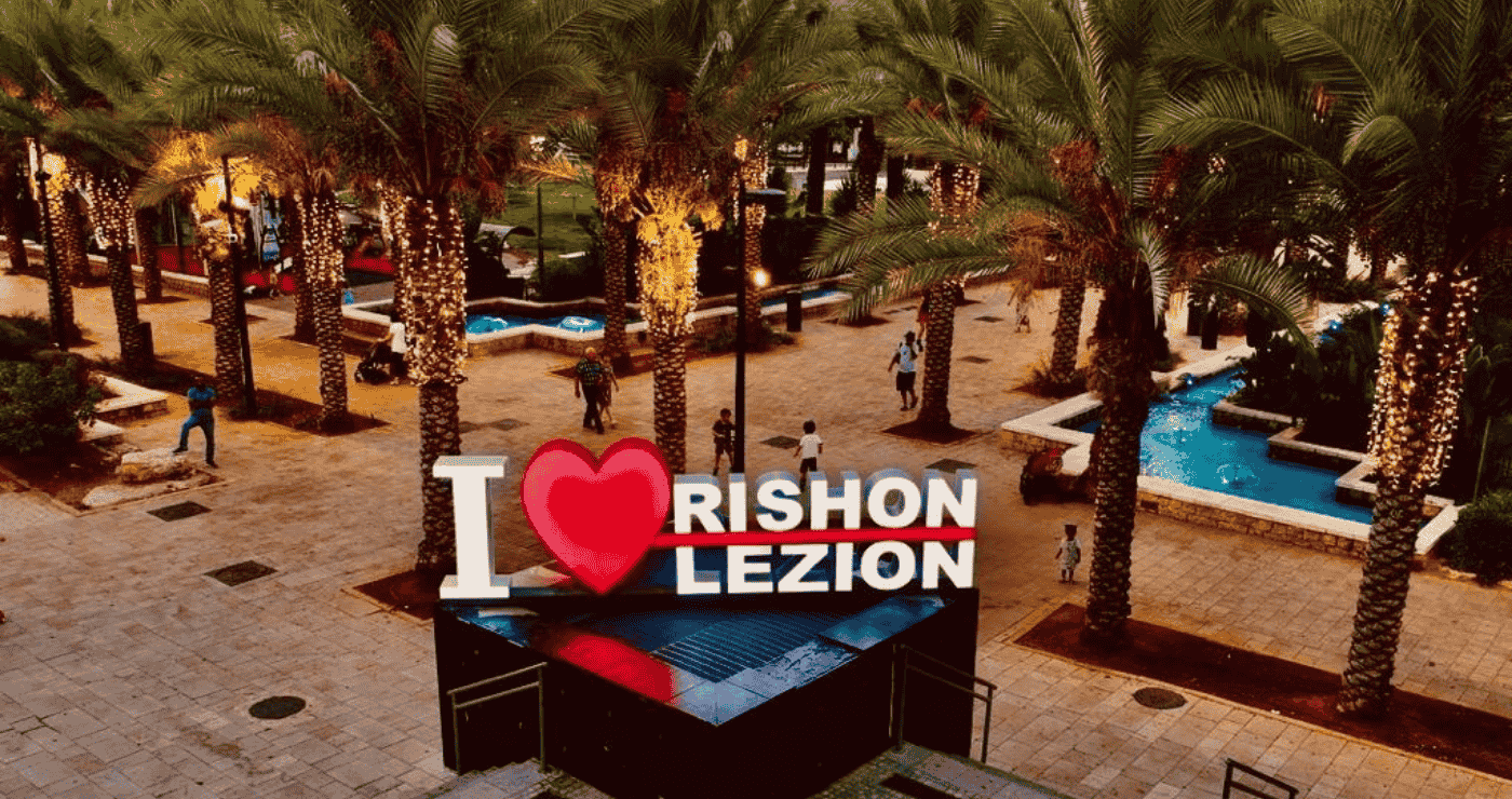 Большая яркая надпись «I ❤️ Rishon Lezion» на освещённой аллее с рядами пальм и небольшими водными бассейнами.