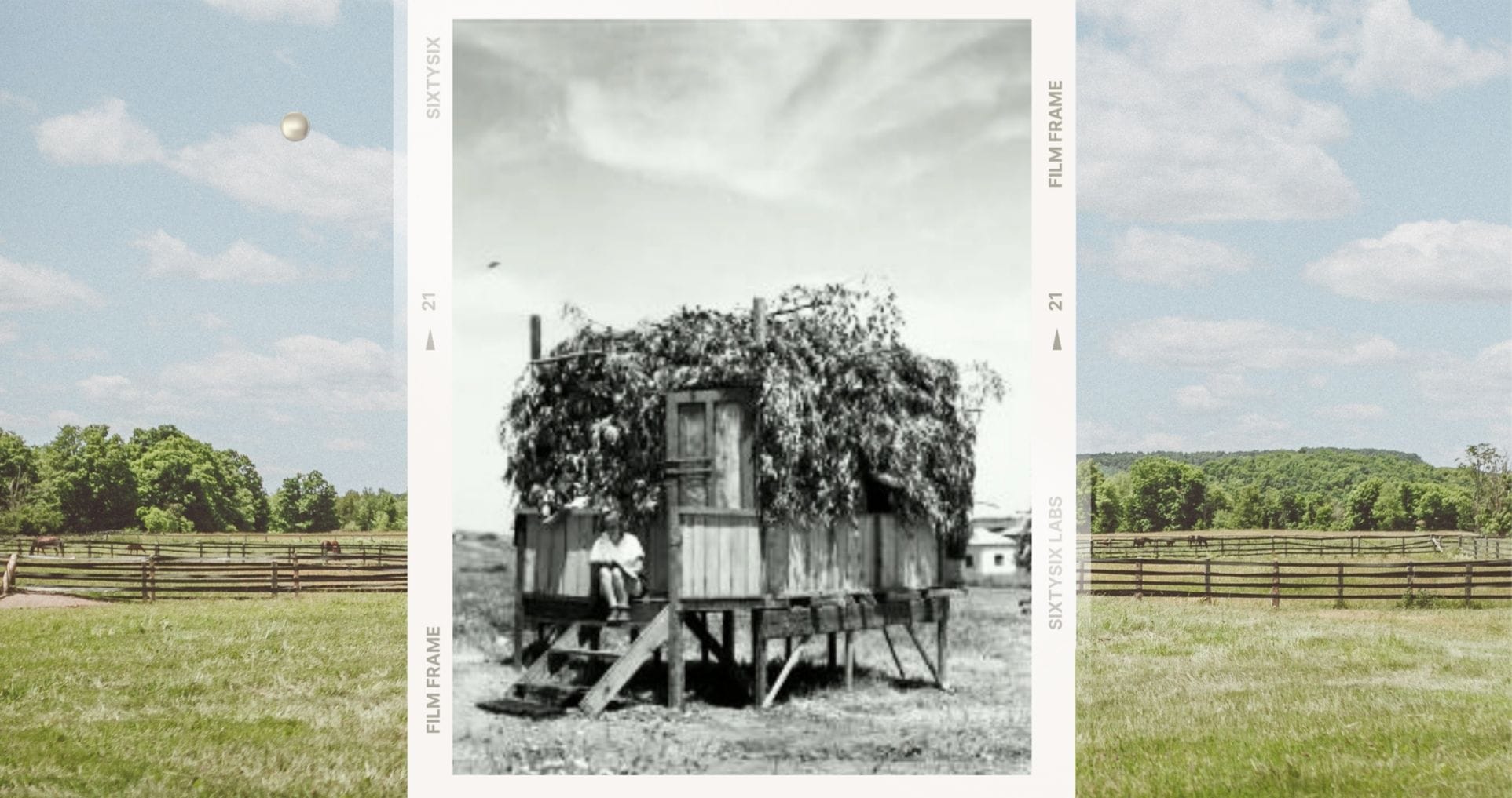Старинная чёрно-белая фотография сукки из ветвей и досок, стоящей на деревянных сваях, в обрамлении современного пейзажа с зелёным полем и забором.