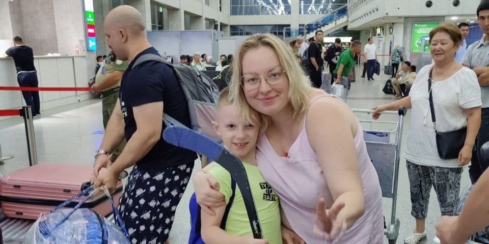 Женщина обнимает мальчика в аэропорту; рядом багаж и люди, ожидающие в зале вылета.